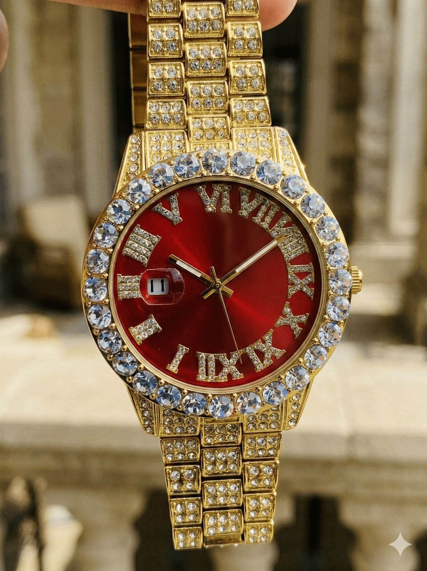 Gold watch with a red face and diamond accents, held against a blurred architectural background.