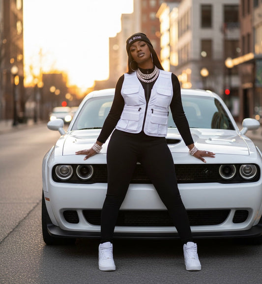 Woman in black jumpsuit, white vest, white air force ones sneakers leans back on white dodge challenger