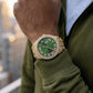 Hand wearing a gold watch with a green face, blurred background