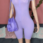 Mannequin wearing a purple bodysuit with a matching handbag against a pink wall.