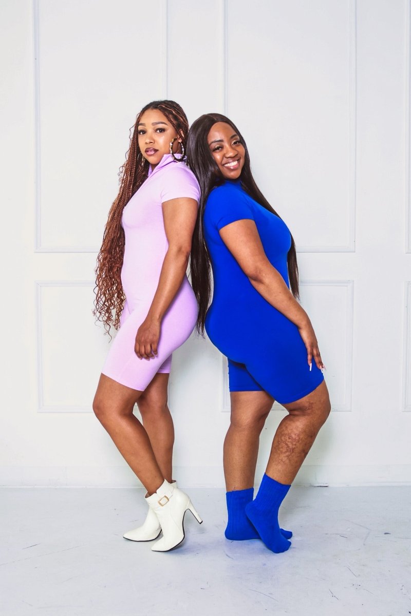 Two women wearing colorful outfits standing back-to-back against a white wall.