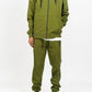 Olive green men’s tech fleece tracksuit, relaxed fit, hands in hoodie pockets, white studio backdrop.