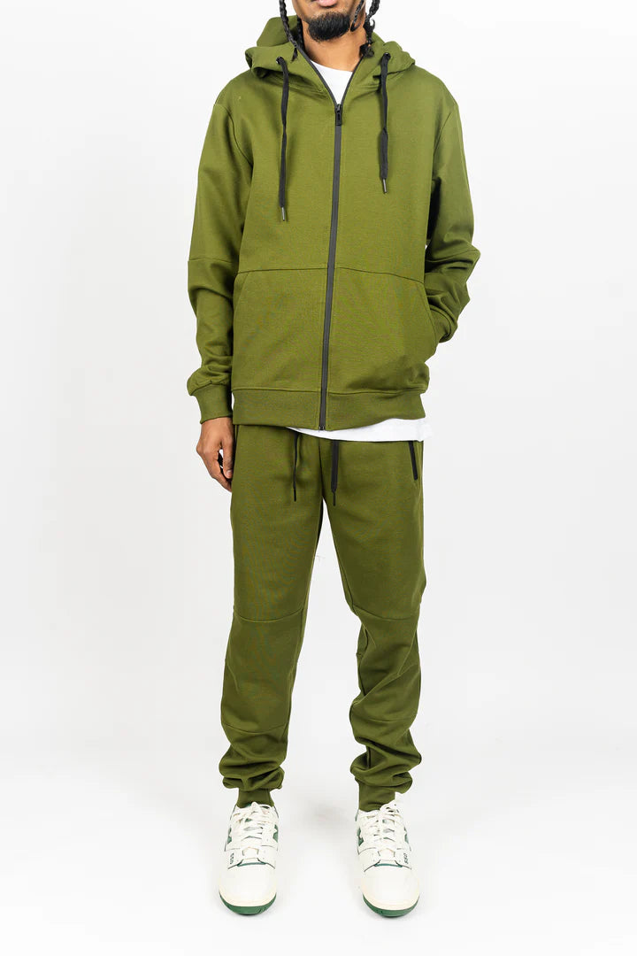 Olive green men’s tech fleece tracksuit, relaxed fit, hands in hoodie pockets, white studio backdrop.