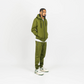 Olive green men’s tech fleece tracksuit, relaxed fit, hands in hoodie pockets, white studio backdrop.
