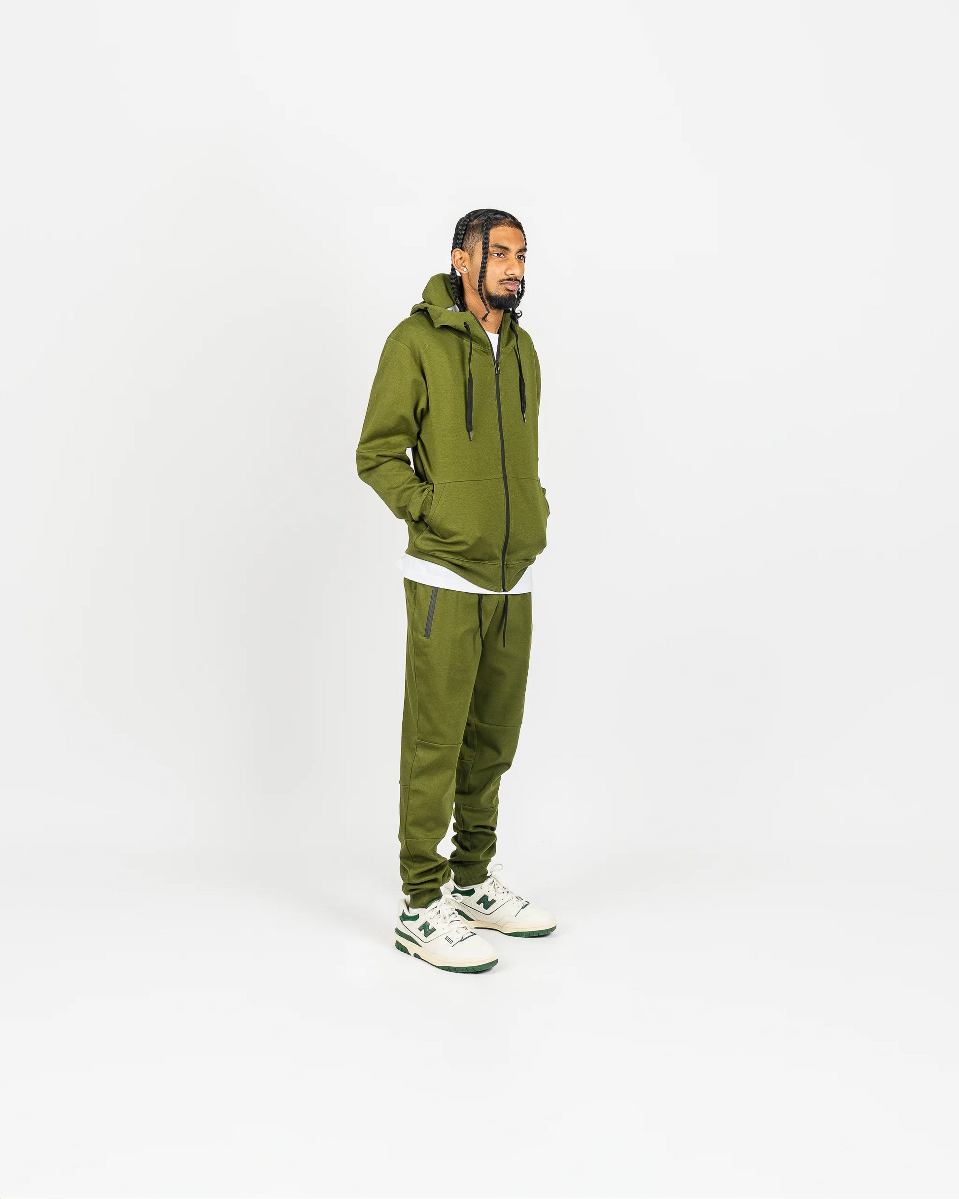 Olive green men’s tech fleece tracksuit, relaxed fit, hands in hoodie pockets, white studio backdrop.
