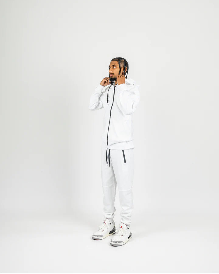 White men’s tech fleece tracksuit with black zipper details, styled with white and red Jordans on white studio background.