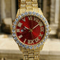 Gold watch with a red face and diamond accents, held against a blurred architectural background.