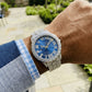 Luxury watch with blue face and diamond accents on a wrist, outdoor background